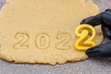 Yeni yıl 2022 Pastanesi konsepti. Siyah eldivenli bir el 2022 tarihini yeni yıl kurabiyeleri ve noel kurabiyeleri için çiğ hamurdan ayırıyor. Mutlu yıllar 2022 konsepti
