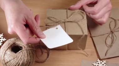 Çevre dostu hediyeler, ev yapımı hediyeler, DIY. Eller, kar taneli ve full HD kaplı bir masada sicim fiyonklu bir kraft karton kutuya boş bir etiket yapıştırıyor. Sıfır Atık Kavramı