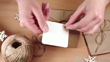 Çevre dostu hediyeler, ev yapımı hediyeler, DIY. Eller boş bir etiketi kar taneli bir masanın üzerinde sicim fiyonklu bir kağıt kutuya yapıştırıyor.