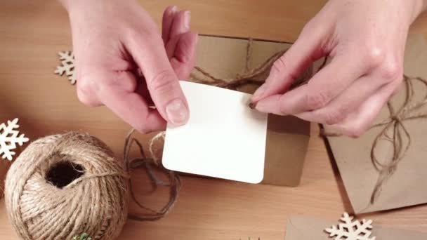 Regalos ecológicos, regalos caseros, bricolaje. Manos pegando una etiqueta en blanco a una caja de papel kraft con un lazo de torzal sobre una mesa con copos de nieve — Vídeos de Stock