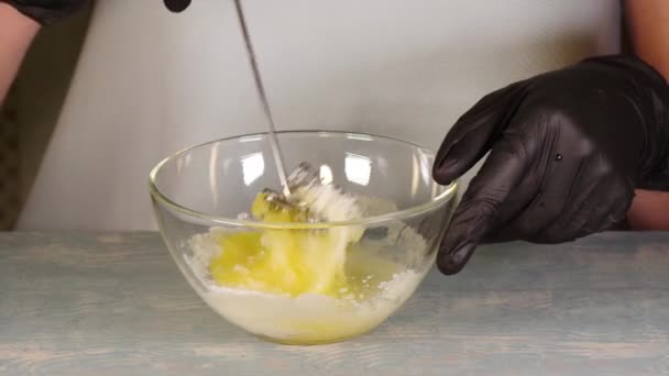 Cozinhar a massa. As mãos em luvas pretas misturam o açúcar e um ovo com um batedor metálico em um boliche transparente, close-up, Full HD 1920 x 1080p — Vídeo de Stock