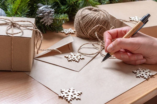 Lista de regalos - Mano sosteniendo un bolígrafo sobre una hoja de papel en blanco, regalos en cajas hechas a mano, copos de nieve y ramas de abeto — Foto de Stock
