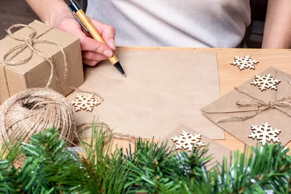 Lista de regalos - Mano sosteniendo un bolígrafo sobre una hoja de papel en blanco, regalos en cajas hechas a mano, copos de nieve y ramas de abeto —  Fotos de Stock