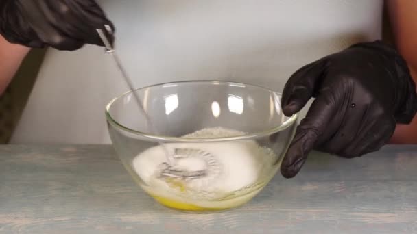 Cozinhar a massa. As mãos em luvas pretas misturam o açúcar e um ovo com um batedor metálico em um boliche transparente, close-up, Full HD 1920 x 1080p — Vídeo de Stock