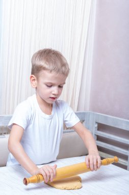 Çocuk mutfaktaki masanın üzerinde merdaneyle hamuru yuvarlıyor. Küçük çocuk annesinin ev yapımı kurabiye pişirmesine yardım ediyor.