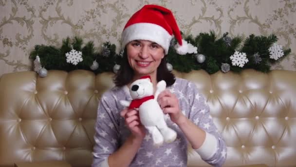 Женщина в красной шляпе Санты и пижаме сидит на кровати, держа плюшевого мишку и танцуя. Счастливого Рождества! — стоковое видео