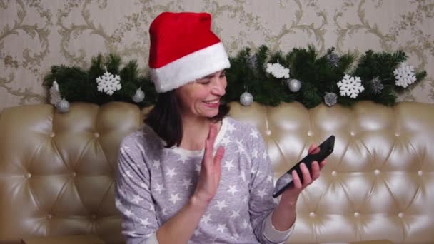 Femme d'âge moyen souriante en pyjama et chapeau de Père Noël ayant un appel vidéo à des amis ou des parents assis sur un lit — Video