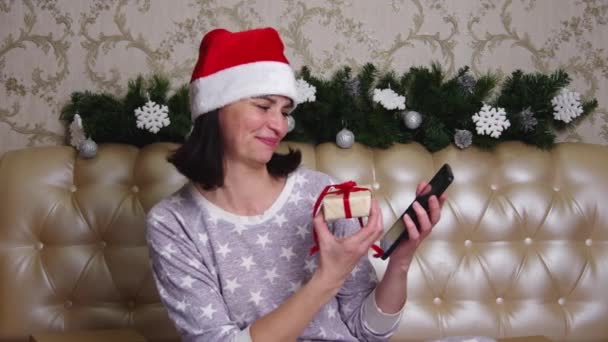 Улыбающаяся женщина в пижаме и шляпе Санты с видеозвонком друзьям, сидящим на кровати, украшенной елкой — стоковое видео