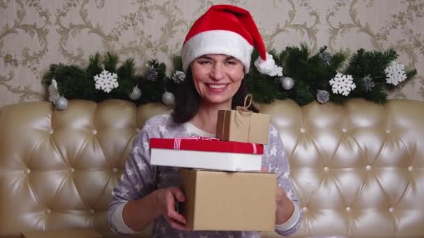 Портрет женщины в красной шляпе Санты и пижаме, сидящей на кровати с несколькими подарочными коробками в руках. Рождественские подарки для мамы — стоковое видео
