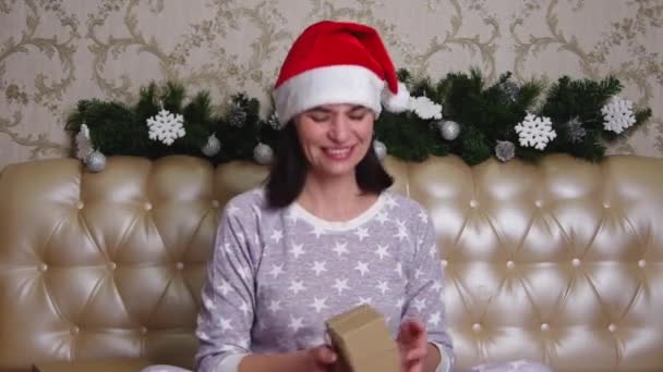 Pijamalı ve Noel Baba şapkalı gülümseyen kadın kusar ve Noel ağacı dallarıyla süslenmiş bir yatağın üzerinde kahverengi bir hediye kutusu yakalar. Kadın aldığı hediyeyle duygusal olarak mutlu. — Stok video