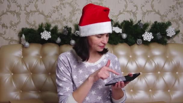 Улыбающаяся женщина средних лет в шляпе Санты использует смартфон дома. Женщина ищет подарки на Рождество и Новый год — стоковое видео