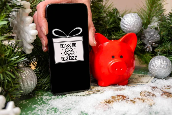 Una mano sosteniendo un teléfono inteligente con un símbolo de regalo y un código QR, una alcancía roja en monedas de oro cubiertas de nieve y árbol de Navidad — Foto de Stock
