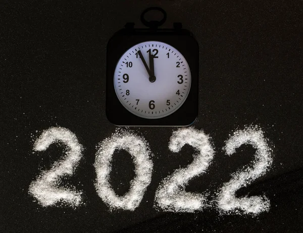 Relógio de alarme preto clássico mostrando o tempo 23: 55 e o ano 2022 escrito com sal branco como neve em um fundo preto. 12 horas. Conceito véspera de Ano Novo. — Fotografia de Stock