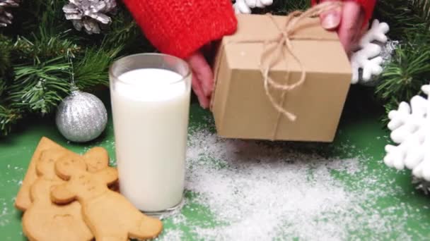 手穿一件红色毛衣，把一个手工礼品盒放在桌子上，桌上有圣诞饼干、一杯牛奶和圣诞装饰品 — 图库视频影像