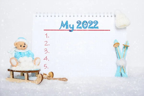 Mavi elbiseli bir oyuncak ayı, mavi ahşap bir kayağın üzerinde oturuyor, beyaz bir şapka ve üzerinde My 2022 yazan bir defter.