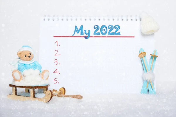 Osito de peluche con ropa azul sentado en un trineo, esquís de madera azul, un sombrero blanco y un cuaderno con la inscripción MY 2022 Fotos de stock
