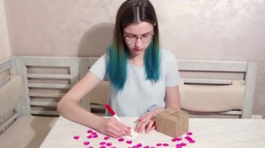 Mavi saçlı ve gözlüklü bir kız, kalbi ve hediyesi olan bir masaya kırmızı bir işaret çiziyor.