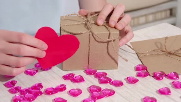 Жіночі руки прикрашають крафтову подарункову коробку з червоним паперовим серцем на столі з рожевими серцями та подарунками . — стокове відео