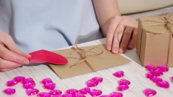 Жіночі руки прикрашають коричневий паперовий конверт, пов'язаний мотузкою з червоним паперовим серцем і показують вам — стокове відео