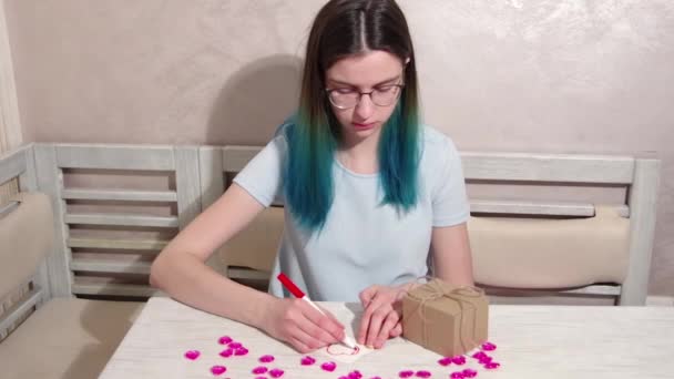 A serious girl with blue hair and glasses decorates a heart shape with a red marker on paper on a table with hearts — Stock video