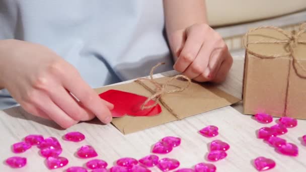 Female hands decorate a brown paper envelope tied with a rope with a red paper heart, full hd, close up. Valentine gift wrapping. Gift for a friend — Wideo stockowe