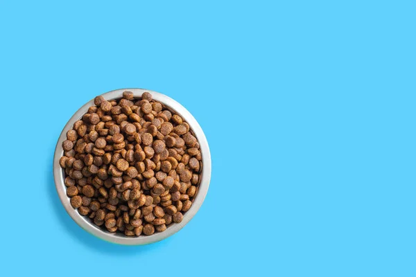 Round metal bowl full of crunchy dry food for dogs or cats on blue background, top view, copy space. Pet care concept — Stock fotografie