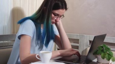 Mavi saçlı gözlüklü yorgun kız dizüstü bilgisayarda yazıyor, kahve içiyor, esniyor. Kız bilgisayarda uyuyakalıyor.