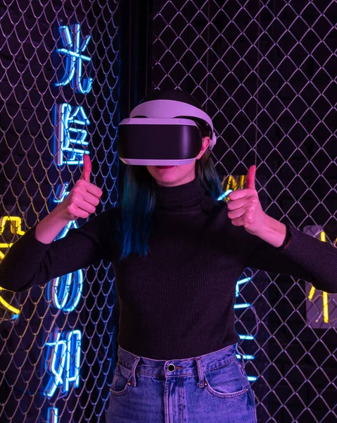 Chica adolescente usando casco vr mostrando pulgares hacia arriba, como gesto sobre fondo colorido neón futurista. Concepto de amor virtual Imagen De Stock