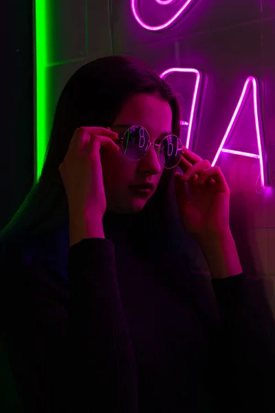 Elegante chica adolescente de moda en gafas con reflejo mira hacia arriba en la calle con iluminación de neón de la ciudad. — Foto de Stock