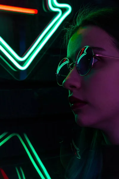 Elegante chica adolescente de moda en gafas con reflejo mira hacia arriba en la calle con iluminación de neón de la ciudad — Foto de Stock