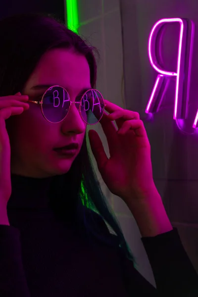 Elegante chica adolescente de moda en gafas con reflejo mira hacia arriba en la calle con iluminación de neón de la ciudad. Concepto de bar y música — Foto de Stock