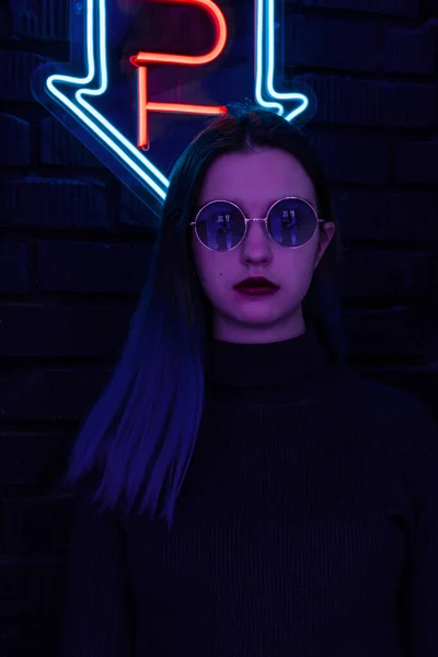 Elegante chica adolescente de moda en gafas con reflejo mira hacia arriba en la calle con iluminación de neón de la ciudad Imagen de archivo