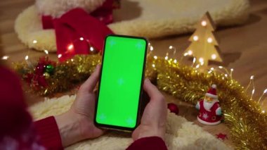 Kadın telefonu yeşil ekranlı tutuyor ve çevrimiçi sohbet ediyor, okuyor, video izliyor. Noel zamanı konsepti. 