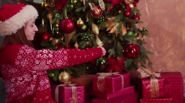 Noel Baba şapkalı genç bir kadın Noel ağacının yanında hediyelerle oturuyor.