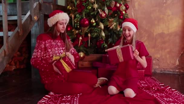 İki mutlu kız kardeş yeni yıl ağacının yanında oturuyor ve hediyeleri açıyor. — Stok video