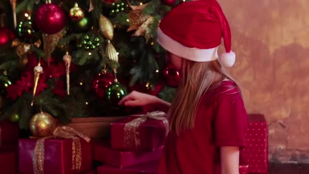 Girl in red santa hat sit near Christmas tree and choose the box with presents — 图库视频影像