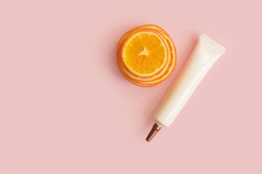 Markasız yüz kremi ve göz kremi uzun başlıklı kozmetik tüp, bronz vida kapağı ve pembe arka planda turuncu dilimler. Model, şablon, kopyalama alanı. C vitaminli krema tüpü..