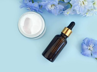 Damgasız kahverengi cam şişede pipetli serum, açık cam kavanozda nemlendirici krem ve mavi arka planda mavi Delphinium çiçeği. Hyaluronik asitle birlikte boş bir kozmetik şişesi. Model, yakın plan..