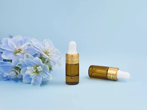 Two gold glass white bottles of hyaluronic acid with pipette and blue Delphinium flower on blue background. Essential oil for care of skin. Mockup. Cosmetic bottles blank with serum.