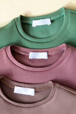 Üç yeni rolive yeşili, tozlu pembe ve bej pamuk gömlekli beyaz etiketler kumaş desenli arka plan. Çocuklar için yuvarlak yakalı uzun kollular.