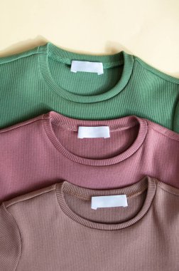 Beyaz boş giysi etiketleri üç yeni rolive green, tozlu pembe ve bej renkli pamuk gömlekler kumaş desenli arka planda. Çocuklar için yuvarlak yakalı uzun kollular.