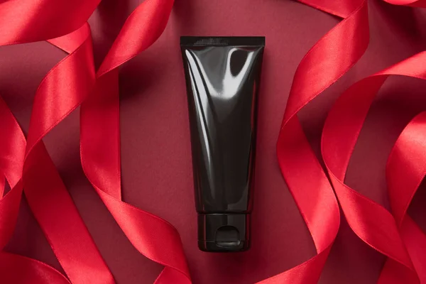 Siyah sıkılmış şişe kozmetik tüp ve kestane rengi arka planda kırmızı kurdeleler. İlaç veya kozmetik ürünleri için tüp - krem, kayganlaştırıcı jel, nemlendirici, cilt bakımı.