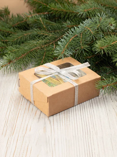 Craft brown paper box with transparent window and a Christmas fir tree on light wooden background. New Year\'s gift box tied with silver ribbon under Christmas fir tree. Vertical photo with copy space.