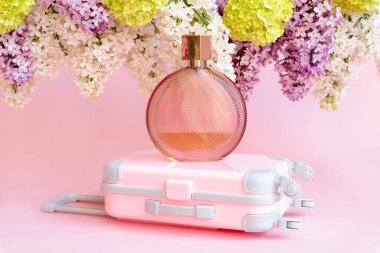 Markasız kahverengi parfüm şişesi, küçük pembe seyahat bavulu oyuncağı, mor ve beyaz leylak ve pembe arka planda yeşil viburnum çiçekleri. Model, şeffaf cam parfüm şişesi markalaştırmak için, ön manzara..