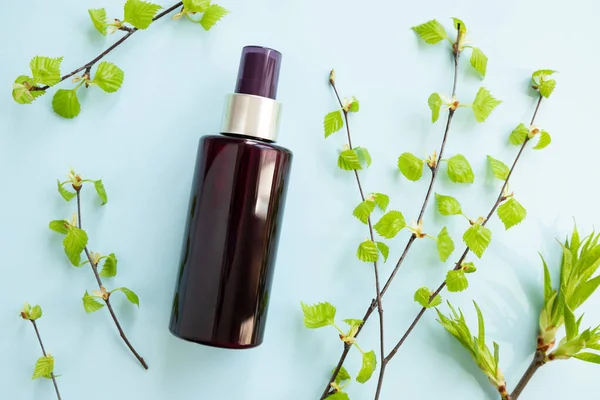 Brown cosmetic spray bottle and birch branches with young small leaves on blue background. Mockup. Skincare beauty and liquid antibacterial spray. Natural Body mist. Front view, flat lay.