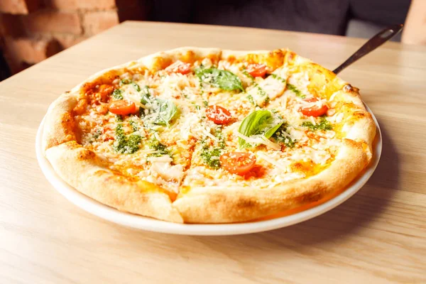 Italian pizza with chicken, cherry tomatoes, mozzarella and parmesan cheese, basil on plate with serving spatula on wooden table. Front view, lifestyle photo, closeup.