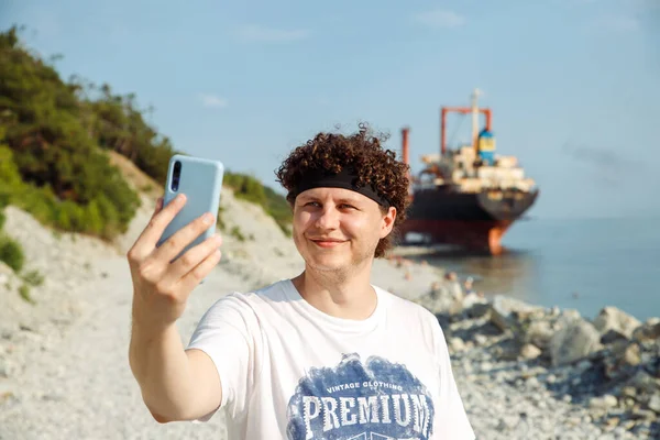 Smiling Young Caucasian man with curly hair making selfie with smartphone. Guy outdoors against sunken cargo ship and taking self portrait with mobile phone. Summer waist up lifestyle portrait