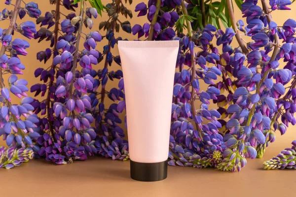 Mockup pink squeeze bottle cosmetic tube with screw black cap and blue purple lupine flowers a lot on brown golden background. Cream, gel, skincare, sunscreen, moisturizer. Front view, blank bottle.