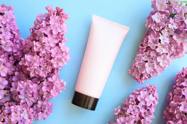 Mockup pink squeeze bottle cosmetic tube with screw black cap and lilac flowers on blue background. Body cream, shower gel, skincare, sunscreen, moisturizer. Front top view, blank tube, flat lay.