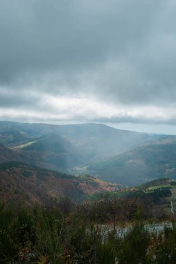 Yeşil, kırmızı, turuncu ve sarı renkli ormanlarla kaplı dağ manzarası manzaraya zıt düşüyor. Mavi ve gri gökyüzü ile doğanın güzel manzarası.